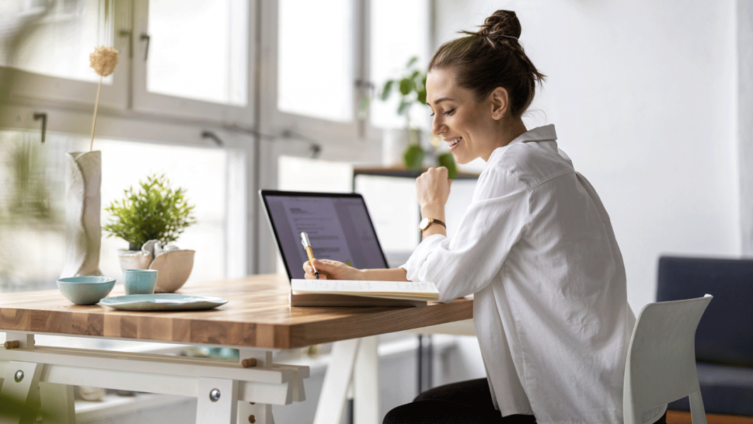 Kvinna som sitter vid ett bord framför laptop och tar anteckningar i anteckningsblock