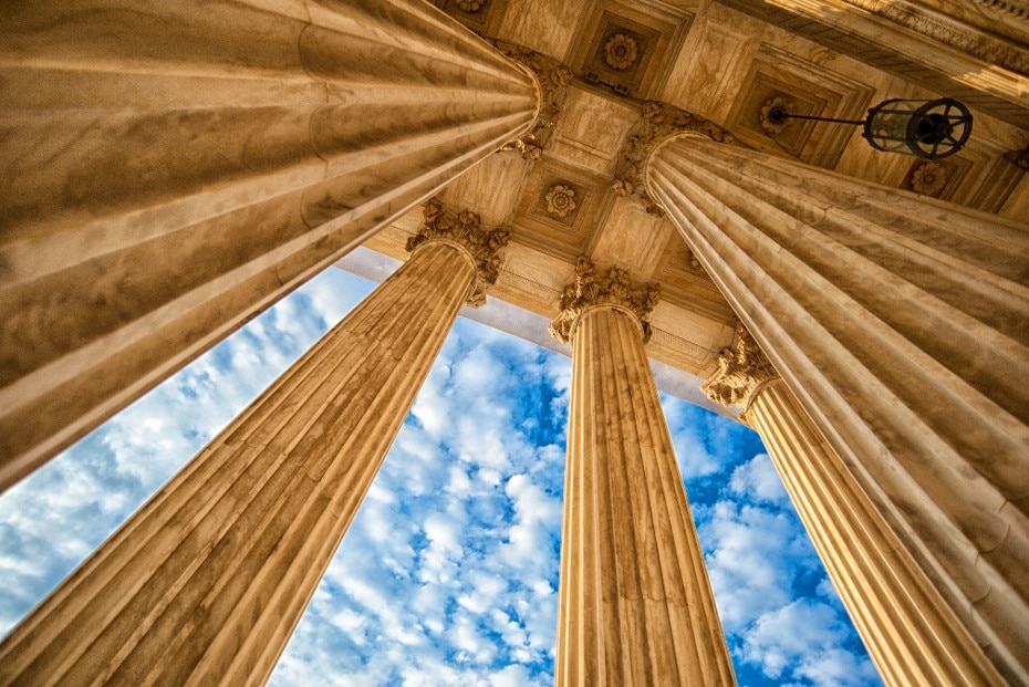 View looking up at tall stone columns and ornate ceiling with blue sky and clouds visible between them