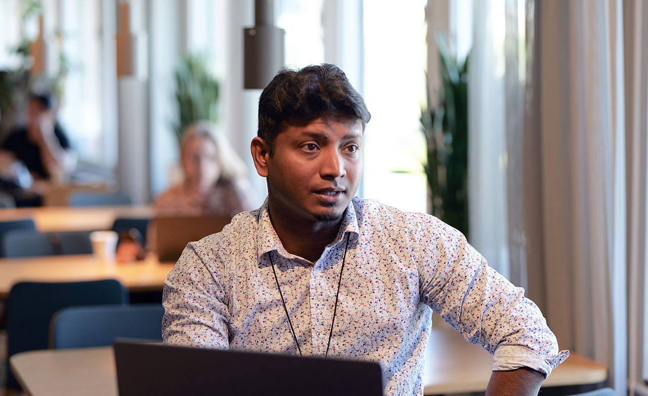 En person sitter vid ett bord med en laptop i en modern kontorsmiljö. Personen är vänd mot någon utanför bild och verkar delta i ett samtal eller möte. I bakgrunden syns andra medarbetare och arbetsplatser i mjukt fokus.