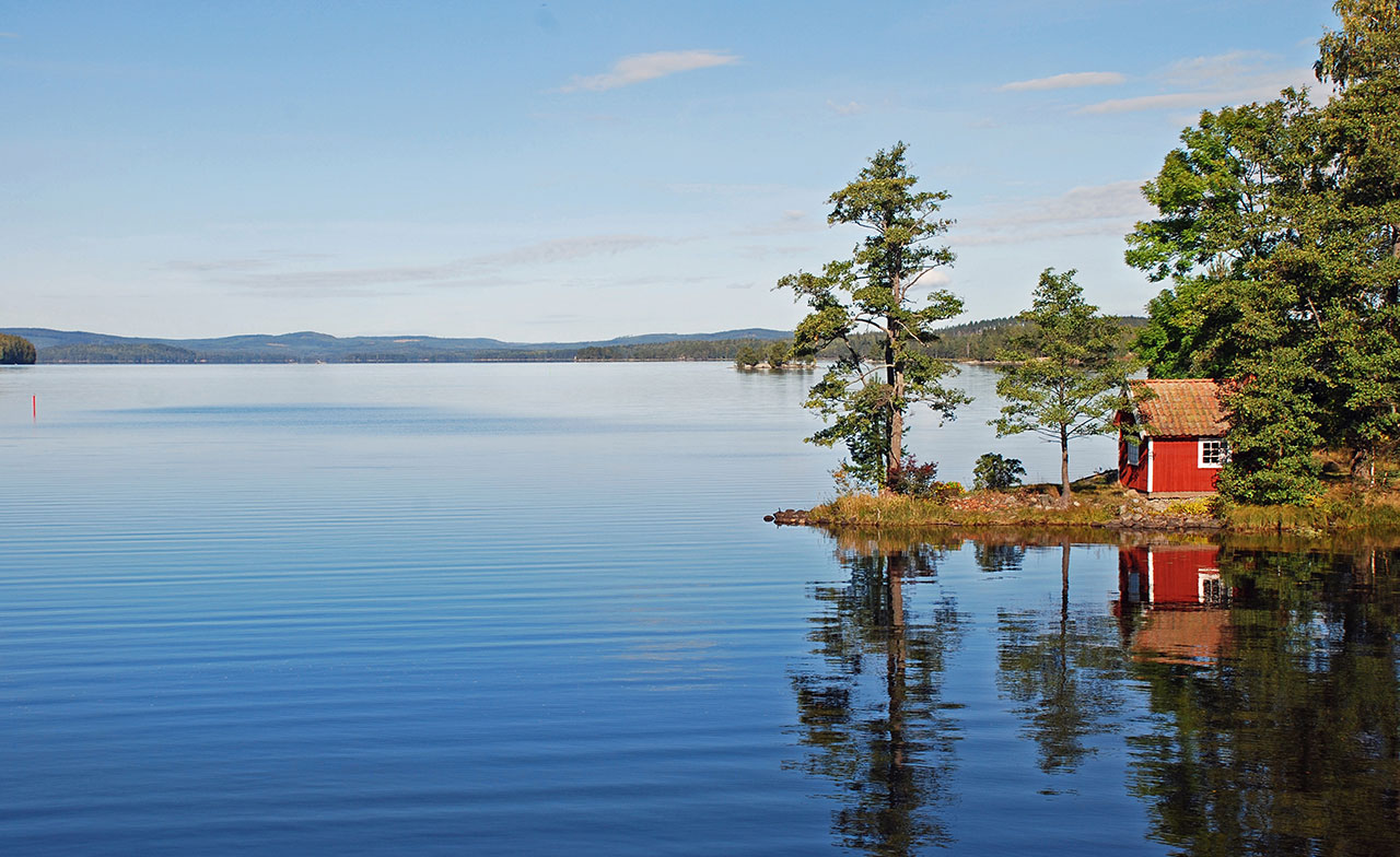 En liten röd stuga ligger vid en stilla sjö omgiven av träd. Vattnet speglar landskapet och himlen är klar, vilket skapar en lugn och naturnära miljö.