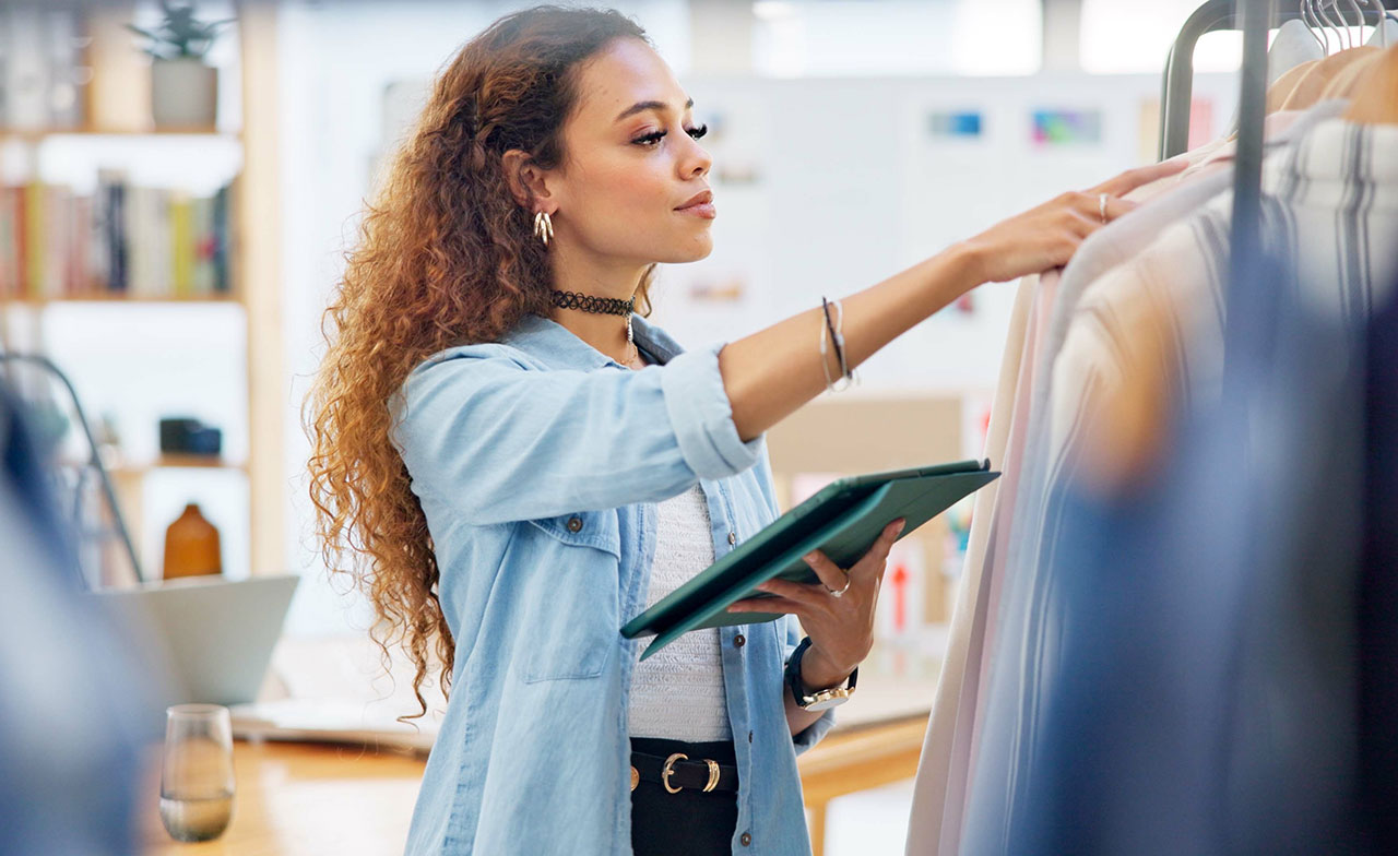 En butikspersonal inspekterar klädesplagg på ett klädställ och håller samtidigt en digital surfplatta. Hon arbetar i en ljus butiksmiljö och verkar kontrollera lager eller prisinformation.