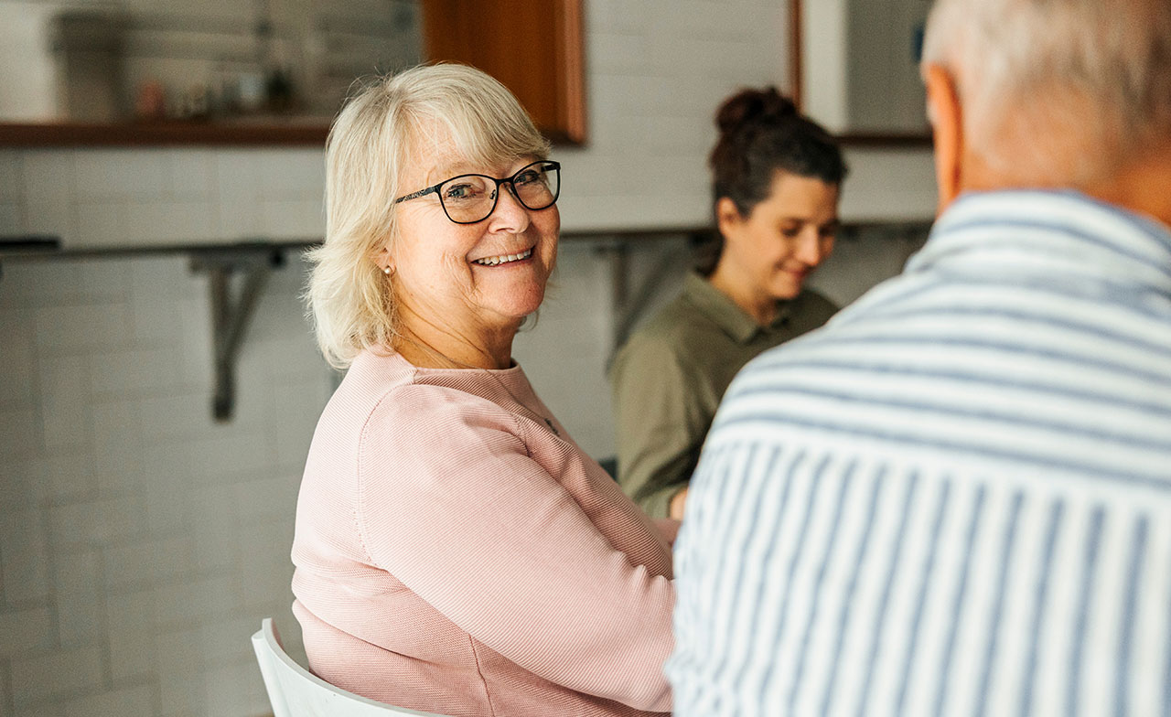En äldre kvinna med glasögon och ljust hår sitter vid ett bord och ler mot någon utanför bild. Andra personer sitter runt henne i ett ljust kök eller vardagsrum, vilket skapar en varm och social stämning.