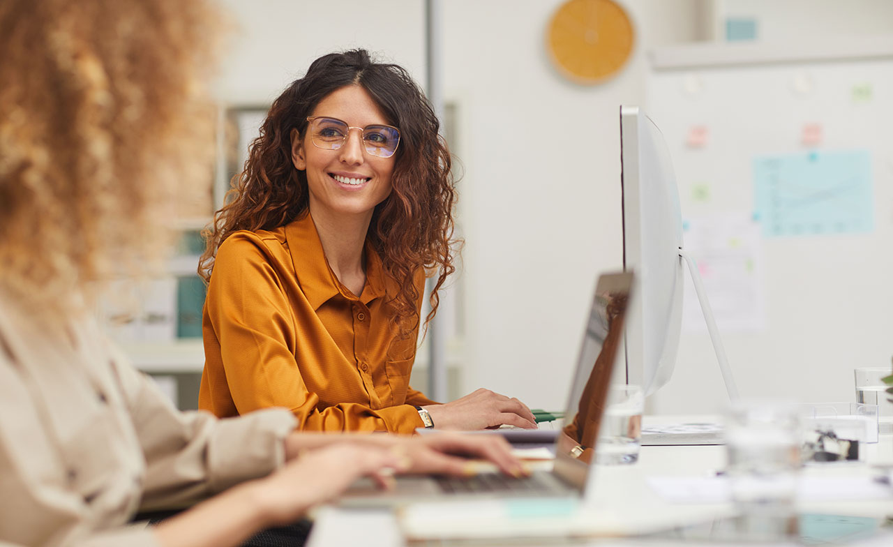 smiling_woman_working_office_colleagues_laptops En leende kvinna med glasögon och orange skjorta sitter vid ett skrivbord på ett kontor, framför en datorskärm. Hon samtalar med en kollega som också arbetar vid en laptop. Kontorsmiljön är ljus och modern.