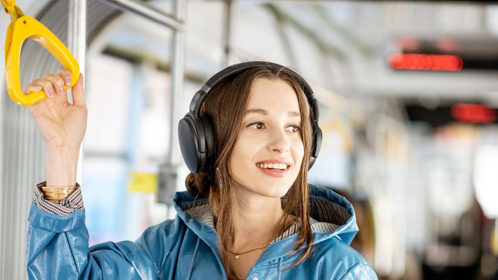 Smiling young female train passenger wearing overear headphones stood holding yellow handle