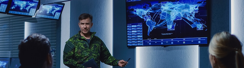 Homme en tenue militaire devant un écran affichant une carte du monde, face à un groupe de personnes Homme en tenue militaire devant un écran affichant une carte du monde, face à un groupe de personnes