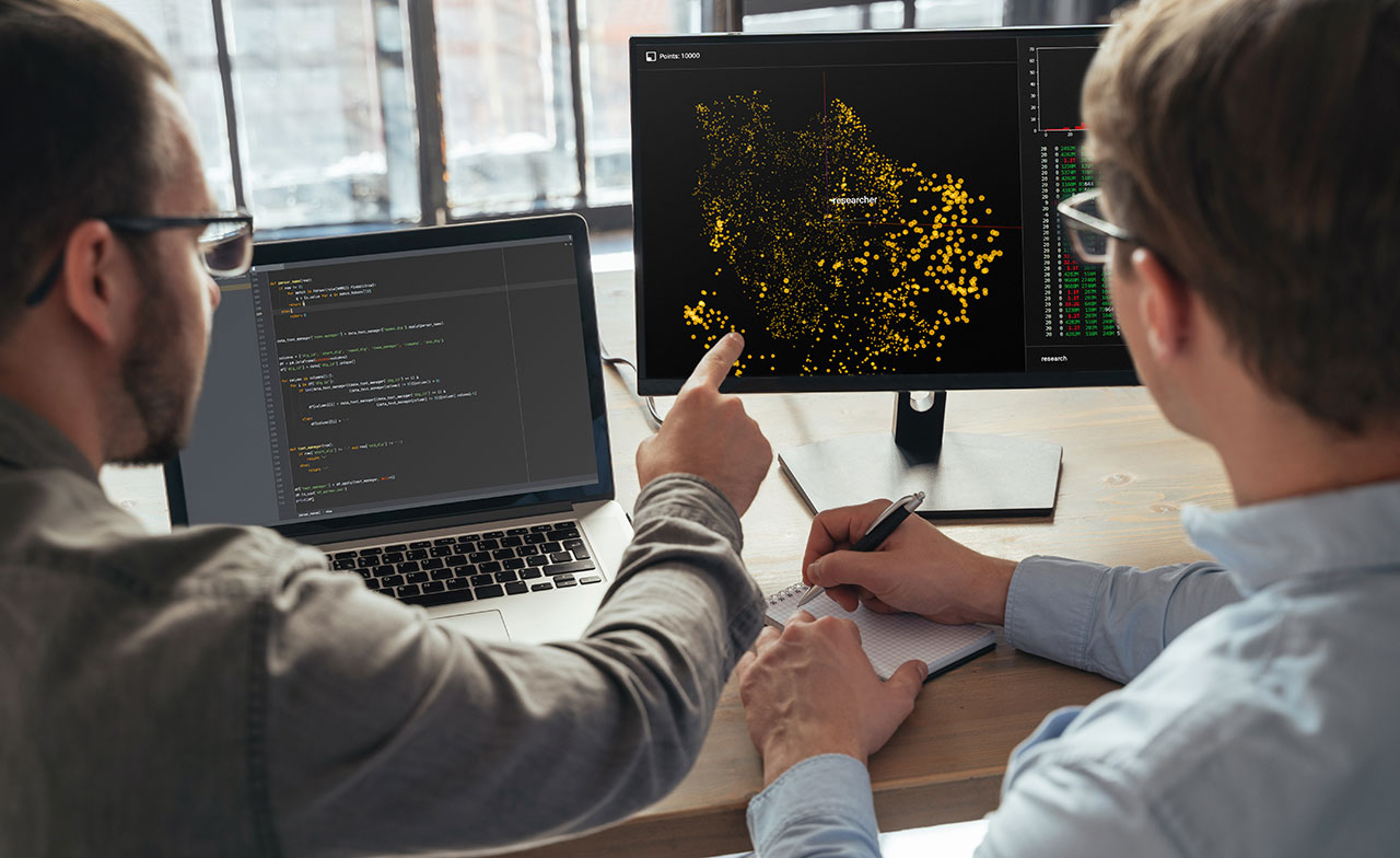 Två personer sitter vid ett skrivbord och analyserar information på två datorskärmar. Den ena pekar på en visualisering med nätverksdata medan kod visas på en bärbar dator bredvid. Bilden illustrerar dataanalys, programmering och tekniskt samarbete.