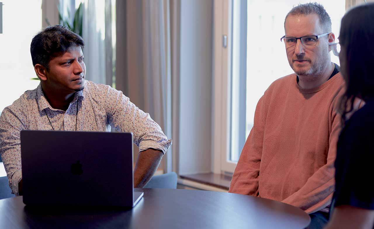 Tre kollegor sitter runt ett bord i en modern kontorsmiljö. En laptop står på bordet medan två av personerna lyssnar uppmärksamt på en tredje person som deltar i samtalet. Stämningen är fokuserad och samarbetsinriktad.
