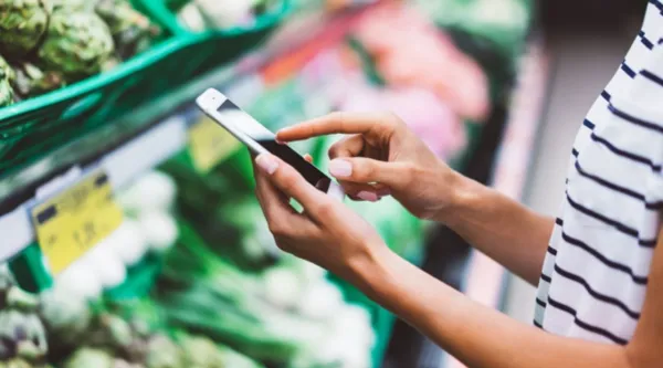 Frau steht im Supermarkt in der Gemüseabteilung und schaut auf ihr Handy