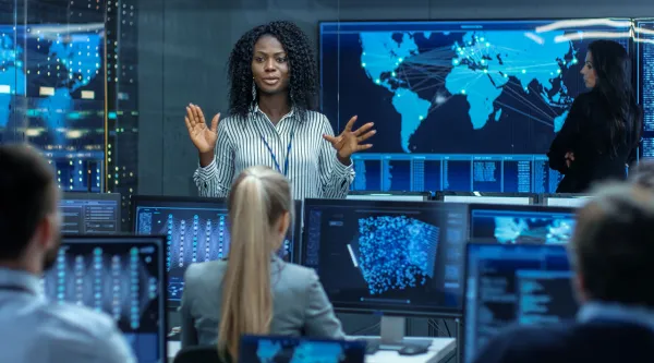 Kvinna håller en presentation i ett kontrollrum med flera skärmar som visar världskartor och dataströmmar, inför en grupp människor.