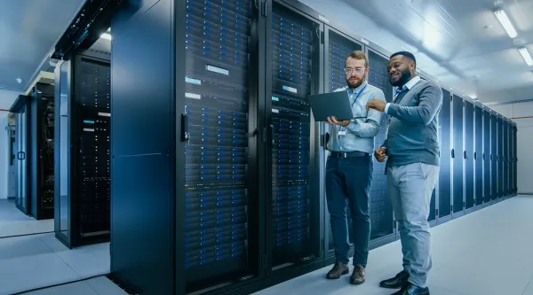 Två IT-tekniker i ett modernt serverrum står framför serverrack och tittar på en bärbar dator medan de diskuterar data eller systemprestanda.