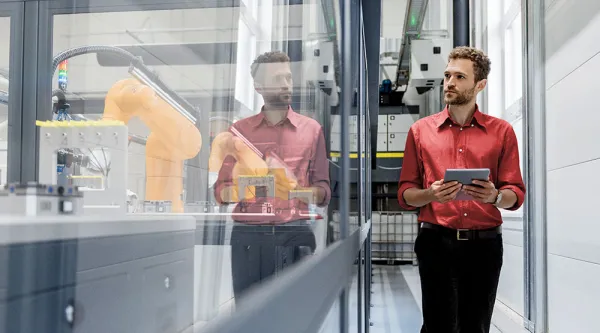 Man i röd skjorta går i industrilokal och tittar in genom fönstret mot robotarm i arbete