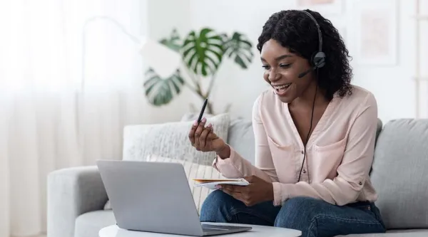 Smiling young woman sitting on sofa and talking on headset while looking at laptop