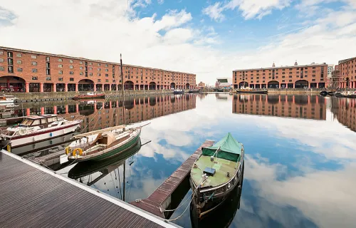 three boats at Albert Docks in Liverpool. The red buildings and cloudy sky are reflected in the…
