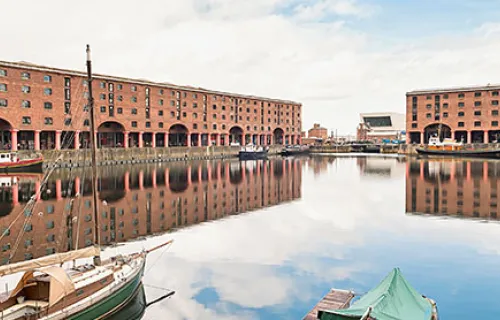  three boats at Albert Docks in Liverpool. The red buildings and cloudy sky are reflected in…