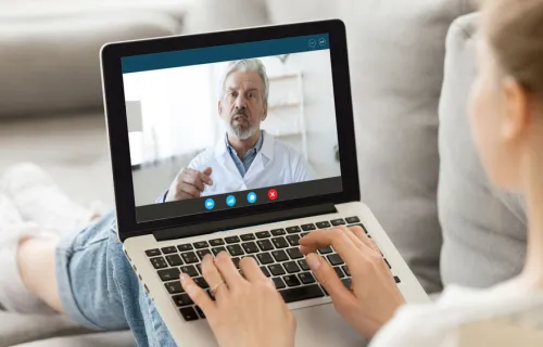 Ung kvinna sitter uppkrupen i sin soffa och har ett videomöte med sin manliga läkare på laptopen