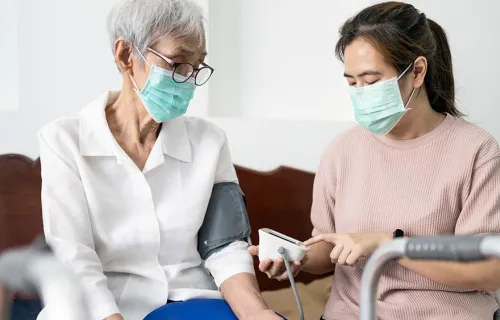 Kvinnlig vårdpersonal med munskydd sitter bredvid äldre kvinnlig patient som också har munskydd…