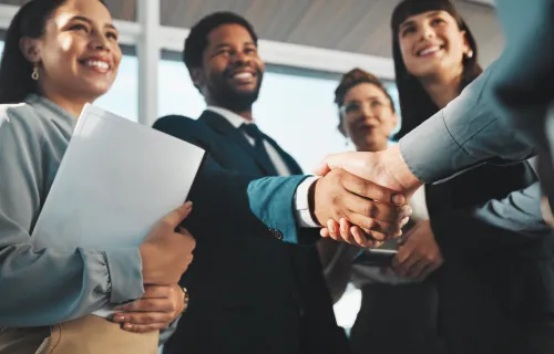 A group of business professionals smiling as two people in the foreground shake hands during a…