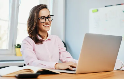 Leende kvinna med glasögon sitter vid arbetsbord och jobbar på sin laptop