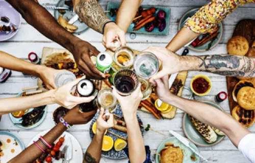 Viele verschiedene Menschen aus unterschiedlichen Nationen stoßen über einem mit Essen belegten Tisch an und symbolisieren so Diversität