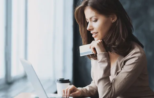 Kvinna sitter vid ett högt cafébord vid ett stort fönster och tittar på sin laptop medan hon håller ett bankkort i vänster hand samtidigt som hon stödjer handen under hakan