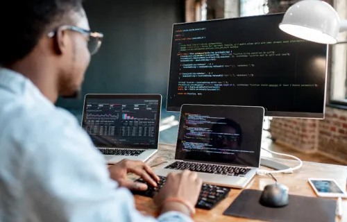 Person working at a desk with two laptops and a large monitor displaying code, data dashboards,…