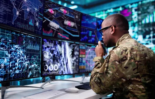 A person in military camouflage uniform sits at a desk with several monitors showing maps,…