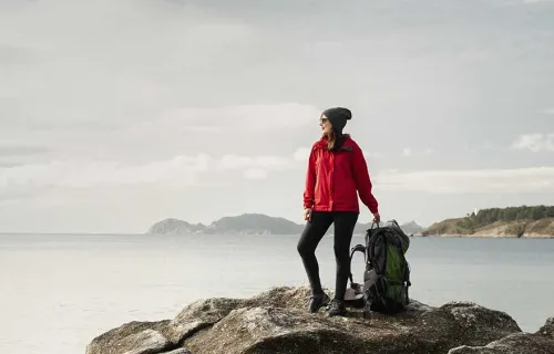Kvinna i röd jacka och mössa står med en stor ryggsäck bredvid sig på en klippa vid havet och…