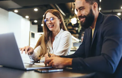 Två kollegor sitter tillsammans vid ett bord och arbetar på en laptop. De ler och diskuterar…