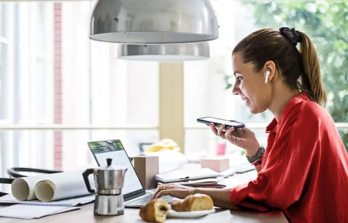 Kvinna i röd topp sitter vid sitt frukostbord i köket och jobbar med laptopen uppfälld och en mobiltelefon i handen