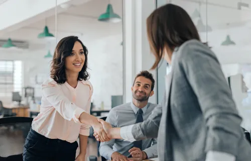 To personer håndhilser i kontorlokale, tredje person ser på og smiler