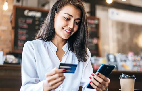 Frau sitzt in einem Cafe und hält in einer Hand ihre Kreditkarte und in der anderen Hand ihr Handy