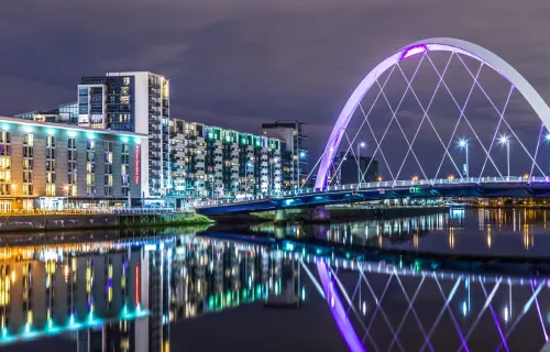 Illuminated white arch bridge spanning a river at night, with modern apartment buildings lining…