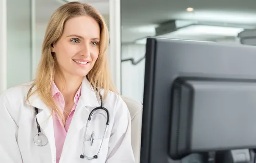 Kvinnlig läkare med vit rock och stetoskop hängande runt halsen tittar leende på sin dataskärm…