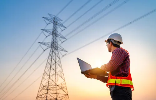 En ingenjör med skyddshjälm och reflexväst står utomhus vid solnedgången och använder en bärbar dator för att inspektera kraftledningar.