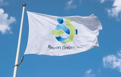 En vit flagga med logotypen för Region Örebro län vajar i vinden mot en klarblå himmel med…