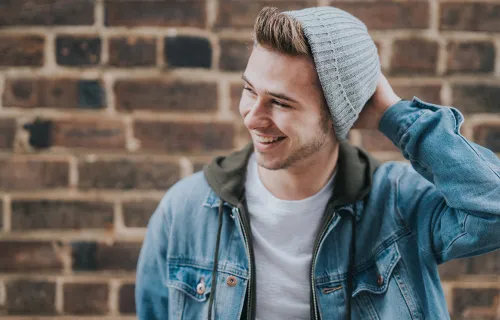 En ung man med jeansjacka och grå mössa står vid en tegelvägg och ler samtidigt som han håller en hand på huvudet och tittar åt sidan