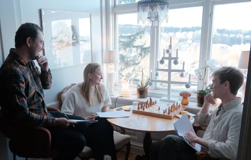 Tre personer sitter vid ett runt bord i ett hemtrevligt rum med stora fönster. De samtalar och…