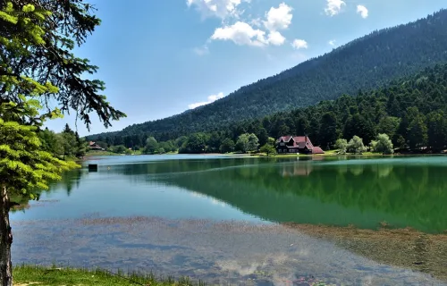 Somrig vy från grässtrand ut över en sjö i en dalgång med träd som klättrar upp för berget på andra sidan och en röd stuga vid stranden