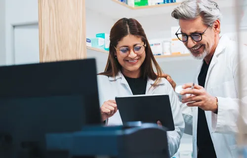 Två vårdmedarbetare i vita arbetskläder står tillsammans och granskar information på en surfplatta. De ler och diskuterar innehållet i en modern vård- eller laboratoriemiljö.