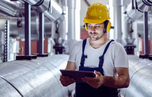 un travailleur d'usine concentré, coiffé d'un casque de protection, utilise une tablette pour vérifier les machines à proximité