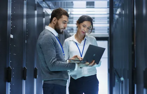 Two people looking at a laptop standing next to server