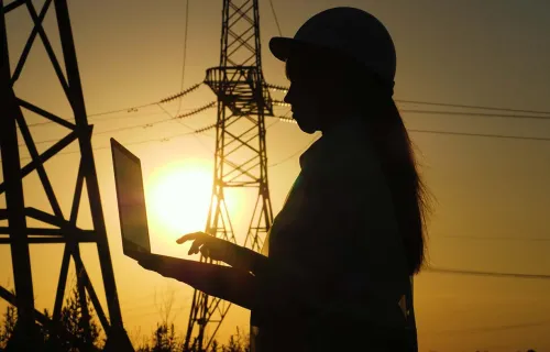 a silhouetted electrical engineer records data on a laptop next to electrical towers at sunset