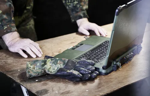 Military personnel using rugged laptop for secure communication on wooden table with camouflage…