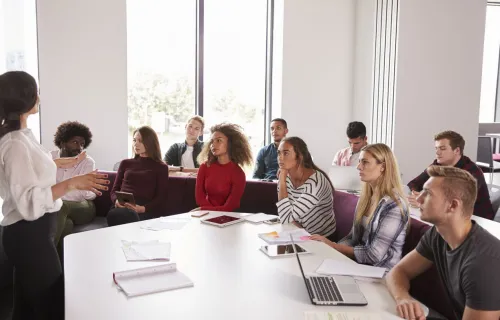 A presenter stands at the front of a meeting room, speaking to a diverse group of students or…