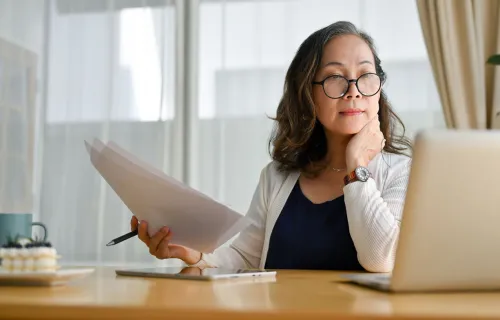 A woman holding paper looking at a laptop screen