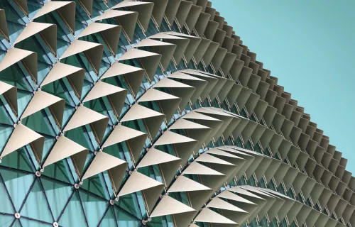 Close-up of a modern glass building facade with rows of white triangular sunshades arranged in…