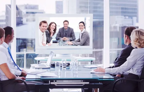 ett antal personer, både kvinnor och män, sitter vid ett konferensbord i ett ljust och luftigt konferensbord för att ha ett möte med kollegor som ansluter på länk och som syns på TV-skärm