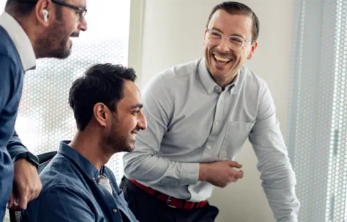 Three men laughing and talking together in a bright office. One man is seated while the other two stand nearby