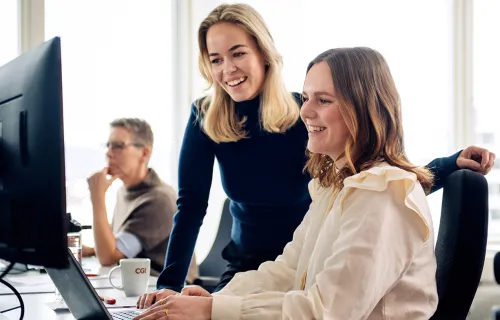 To personer smiler foran dataskjerm, tredje sitter i bakgrunnen.