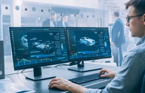 Männlicher Datenwissenschaftler betrachtet zwei Computerbildschirme in einer Autofabrik.